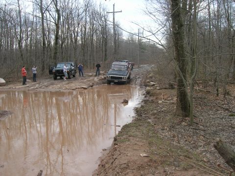 1996 Ford Bronco - A group of people stand around several off-road vehicles stuck in a large mud puddle on a wooded trail. One black SUV is partially submerge…