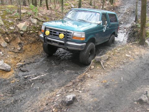 1996 Ford Bronco - A teal 1996 Ford Bronco is navigating a muddy, rocky trail in a wooded area. The vehicle is angled towards the viewer's left, with its fron…
