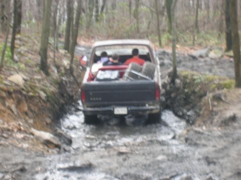1996 Ford Bronco - A dark-colored pickup truck drives through a muddy, wooded trail. The truck has three people visible in the cab and bed, and appears to be…