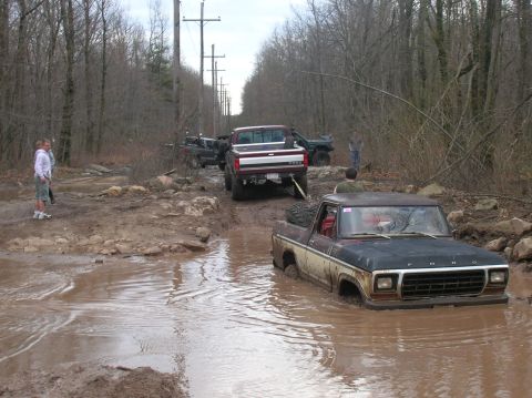 1996 Ford Bronco - Two vintage Ford pickup trucks are stuck in deep mud on a wooded trail. A maroon and white pickup is partially submerged in the foreground,…