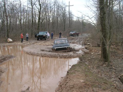1996 Ford Bronco - A dark-colored SUV is stuck in a deep mud puddle on a dirt road surrounded by trees. Several people are standing around watching as another…