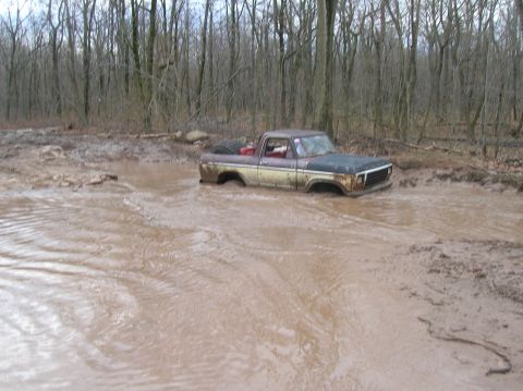 1996 Ford Bronco - A rusty, two-tone pickup truck is stuck deep in a muddy forest puddle. The truck appears to be a classic model with a brown and tan exterio…