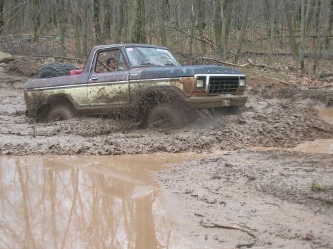 1996 Ford Bronco - A vintage Ford Bronco is shown driving through a deep mud pit, with mud splashing high around the vehicle. The truck appears to be a two-to…