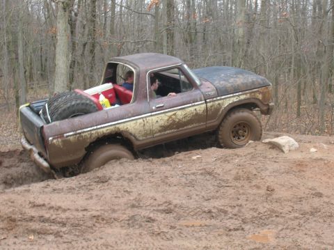 1996 Ford Bronco - A rusty, mud-covered 1970s era pickup truck is stuck in deep mud in a wooded area. The truck has a spare tire mounted in the bed and two pe…