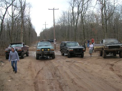 1996 Ford Bronco - A group of off-road vehicles are parked on a muddy dirt road lined with trees. A woman in a grey hoodie walks towards the camera in the for…