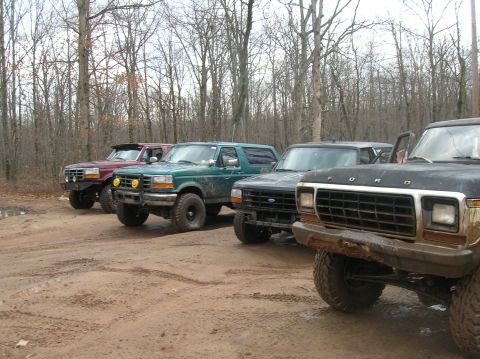 1996 Ford Bronco - Four off-road modified Ford trucks and SUVs are parked in a line on a dirt road in a wooded area. The closest vehicle is a dark-colored For…