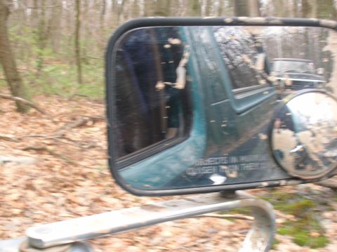 1996 Ford Bronco - A side mirror reflects a forest scene and another vehicle behind the current one, which is partially visible and covered in mud. Text is vi…