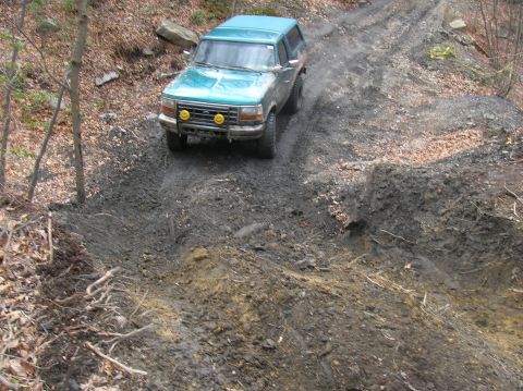 1996 Ford Bronco - A teal Ford Bronco is navigating a muddy, off-road trail. The vehicle is angled downhill with its front facing the camera, showing two yell…