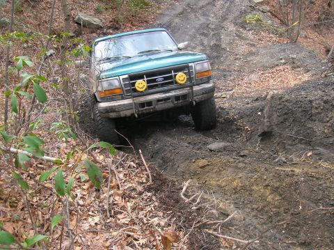 1996 Ford Bronco - A green Ford Bronco is navigating a muddy, off-road trail. The vehicle is tilted to the side as it drives through a rutted path surrounded…