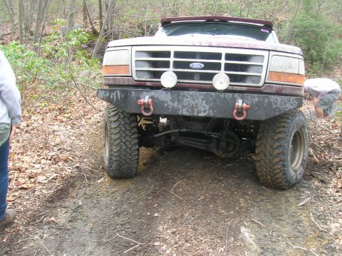 1996 Ford Bronco - The front view of a dark red Ford Bronco is shown off-road, with large mud tires and a custom bumper. A person is visible to the left, and…