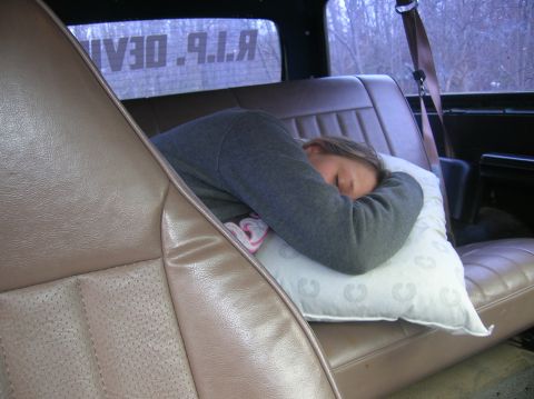 1996 Ford Bronco - A person sleeps on a pillow in the back seat of a vehicle, with a seatbelt visible and text reading "R.I.P. DEAN" on the rear window.