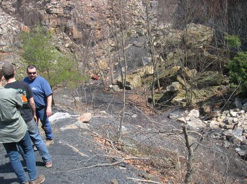 1996 Ford Bronco - Three people stand in a rocky, wooded area with a steep cliff face in the background. The ground is covered in dark gravel and scattered ro…