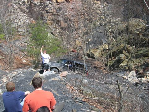 1996 Ford Bronco - A man in a white shirt stands next to a dark-colored SUV that is partially submerged in a rocky, wooded area. Two men are sitting in the fo…