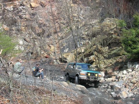 1996 Ford Bronco - A teal and silver Ford Bronco is parked on a rocky, wooded trail. Two people are sitting on the ground to the left of the vehicle.