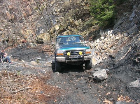 1996 Ford Bronco - A teal Ford Bronco is driving on a rocky, dirt path with trees and a rocky embankment in the background. Two yellow smiley face decals are…