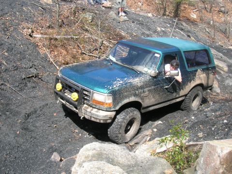 1996 Ford Bronco - A muddy, teal-green Ford Bronco is navigating a rocky, off-road trail. The vehicle has two yellow smiley face fog lights mounted on the fro…