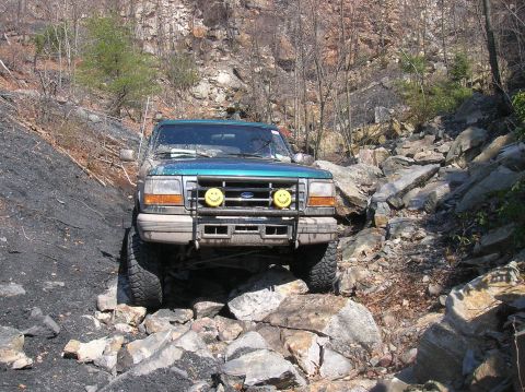 1996 Ford Bronco - A teal Ford Bronco is navigating a rocky, off-road trail. The front of the SUV is visible, equipped with a brush guard and two yellow fog l…
