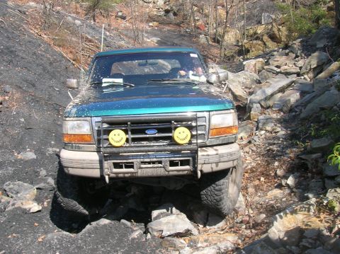 1996 Ford Bronco - A teal 1996 Ford Bronco is navigating a rocky, off-road trail. The vehicle is equipped with a brush guard and two yellow auxiliary lights m…