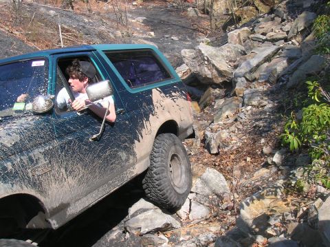 1996 Ford Bronco - A dark green Ford Bronco is navigating a rocky, off-road trail, with mud splattered across its body. The driver, wearing a black baseball c…