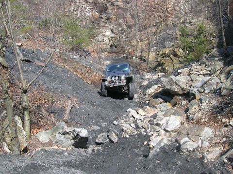 1996 Ford Bronco - A blue Jeep Wrangler is driving up a steep, rocky, and muddy incline in a quarry. The vehicle is angled towards the viewer as it navigates…
