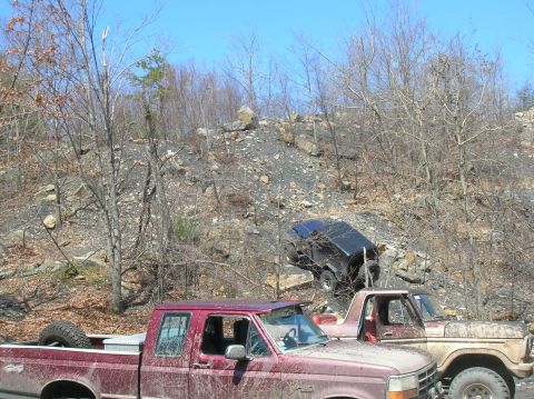 1996 Ford Bronco - A red Ford pickup truck is parked in the foreground, covered in mud. In the background, a black Jeep is parked precariously on a rocky incl…