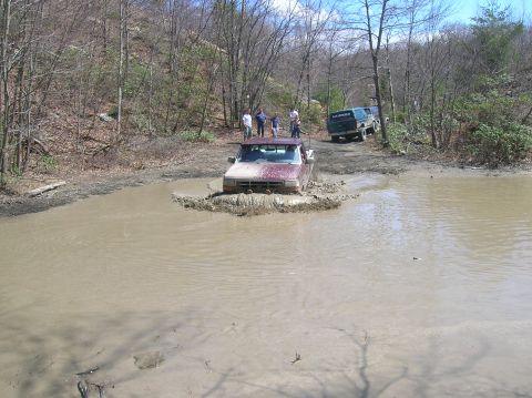 1996 Ford Bronco - A maroon pickup truck is driving through a muddy puddle, kicking up a large splash. Several people stand on the bank watching.