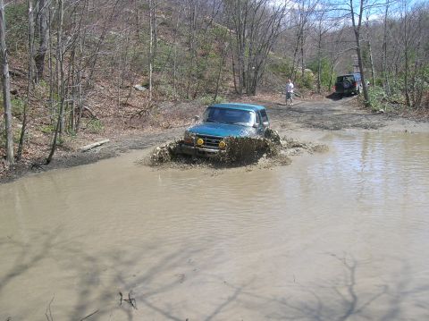 1996 Ford Bronco - A teal Ford Bronco is driving through a muddy puddle, kicking up a large splash. Another dark colored SUV is parked on a dirt road in the b…