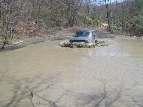 1996 Ford Bronco - A dark-colored SUV is driving through a deep mud puddle, creating a large splash. The vehicle is surrounded by trees and a dirt road.
