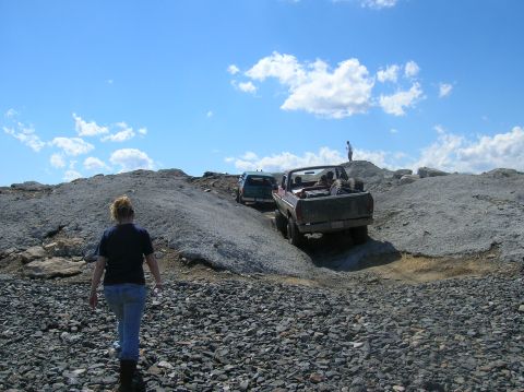 1996 Ford Bronco - A dark red Ford pickup truck is driving up a rocky incline under a blue sky with scattered clouds. A person is standing on a rocky outcrop…