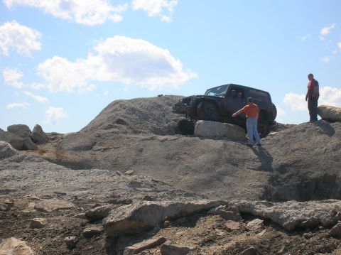 1996 Ford Bronco - A dark-colored Jeep Wrangler is positioned on a rocky incline, with two men standing nearby, one appearing to guide the vehicle.