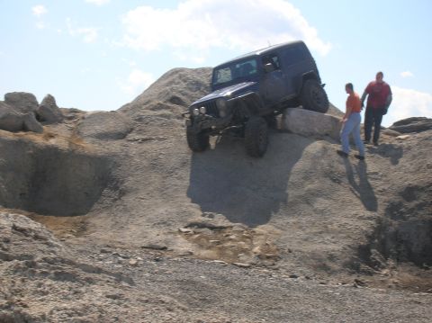 1996 Ford Bronco - A dark blue Jeep Wrangler is navigating a steep, rocky incline with two people standing on the rocks nearby. The Jeep is tilted at an angle…