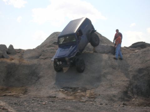 1996 Ford Bronco - A dark blue Jeep Wrangler is angled precariously on a rocky incline, with its front wheels elevated and its rear wheels on the ground. Two…