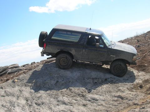 1996 Ford Bronco - A black Ford Bronco is shown driving up a rocky incline under a blue sky with scattered clouds. The vehicle has a spare tire mounted on the…