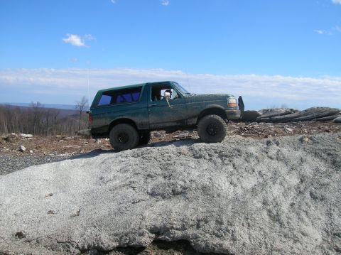 1996 Ford Bronco - A dark green, lifted Ford Bronco is parked on a rocky outcrop under a bright blue sky with scattered clouds. The vehicle is angled towards…