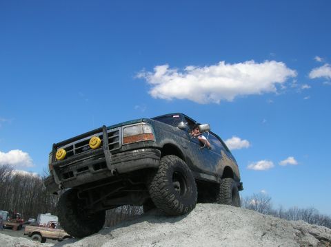 1996 Ford Bronco - A dark green, lifted Ford Bronco is shown from a low angle, climbing a dirt mound under a bright blue sky with scattered clouds. The vehicl…