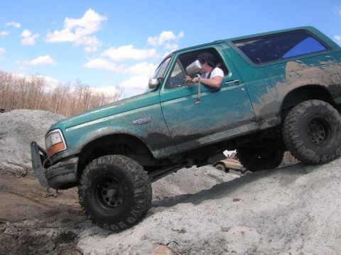 1996 Ford Bronco - A teal Ford Bronco is shown at an angle, climbing over a rocky, sandy terrain under a cloudy blue sky. The vehicle is heavily muddied, indi…