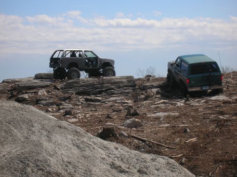 1996 Ford Bronco - Two modified off-road SUVs are parked on a rocky, uneven terrain under a partly cloudy blue sky. The vehicle on the left is a gray SUV with…