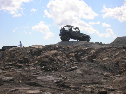1996 Ford Bronco - A dark green Jeep Wrangler with a roll cage is parked on a rocky incline under a blue sky with scattered clouds. A person in a white shirt…