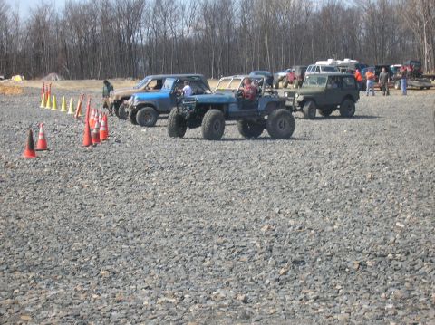 1996 Ford Bronco - Several off-road vehicles are parked on a gravel lot with trees in the background. A blue Jeep-like vehicle with large tires and a roll cag…
