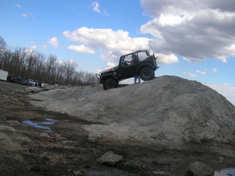 1996 Ford Bronco - A black Jeep Wrangler is climbing a steep dirt and rock hill under a cloudy blue sky. Several other vehicles are parked in the background n…