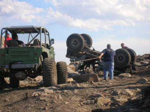 1996 Ford Bronco - Two off-road vehicles are on a rocky terrain under a cloudy sky. One green vehicle is upright, while another vehicle is overturned with two…
