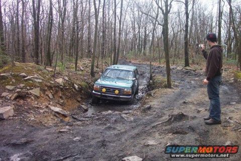1996 Ford Bronco - A teal Ford Bronco is navigating a muddy, wooded trail. A person stands to the right, holding up a phone to take a picture of the vehicle.