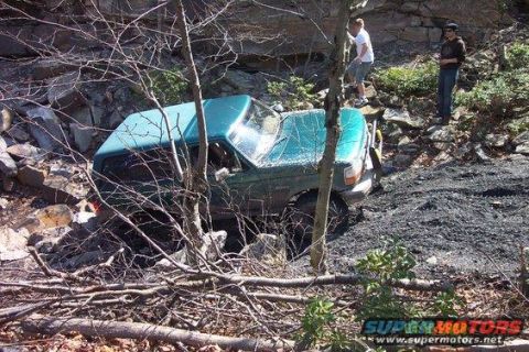 1996 Ford Bronco - A teal Ford Bronco is partially submerged in a rocky, wooded ravine with two people observing from the embankment above. The vehicle appear…