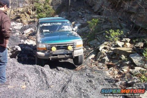1996 Ford Bronco - A teal Ford Bronco is navigating a rocky, off-road trail. The vehicle has two yellow smiley face lights mounted on the front brush guard.