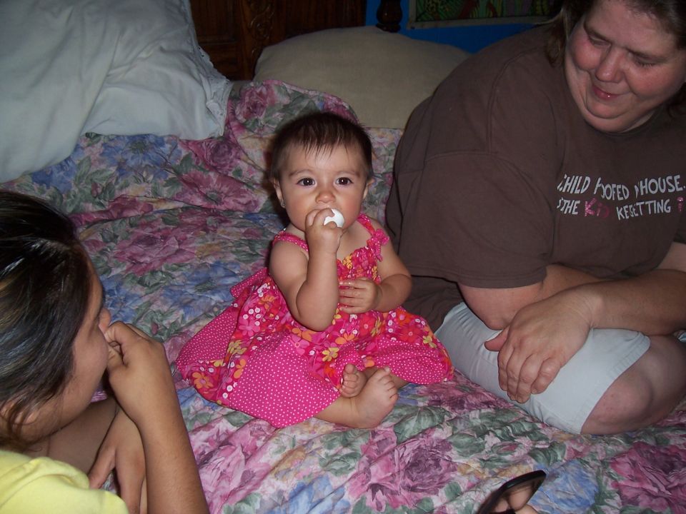 Baby in a pink dress sits on a floral bedspread with two adults