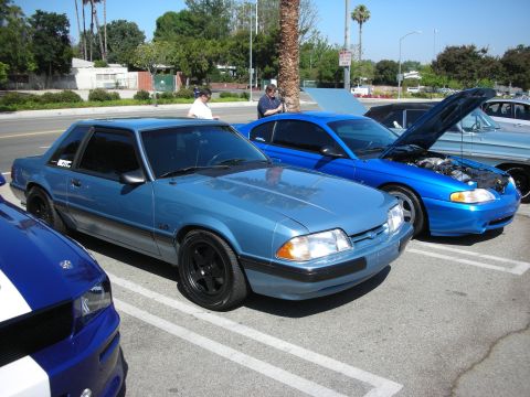 1990 Ford Mustang - Three cars are parked in a lot on a sunny day. The blue coupe in the foreground has black wheels.
