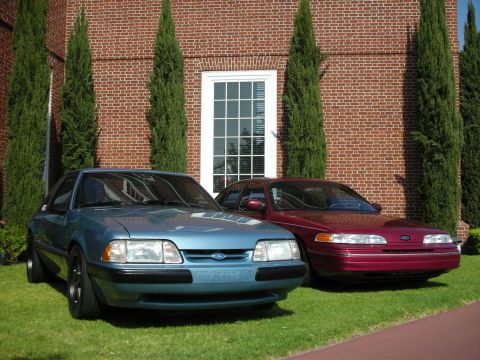 1990 Ford Mustang - Two Ford sedans are parked on a lawn in front of a brick building with a large window.