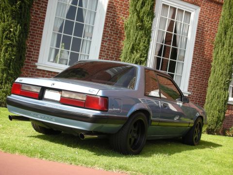 1990 Ford Mustang - Gray Fox-body Ford Mustang LX coupe with black wheels parked on grass before a brick building, rear view.
