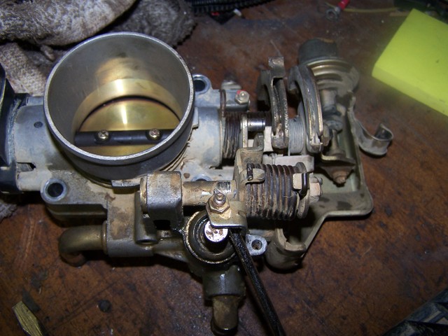 Removed automotive throttle body with brass bore and linkage springs on a workbench