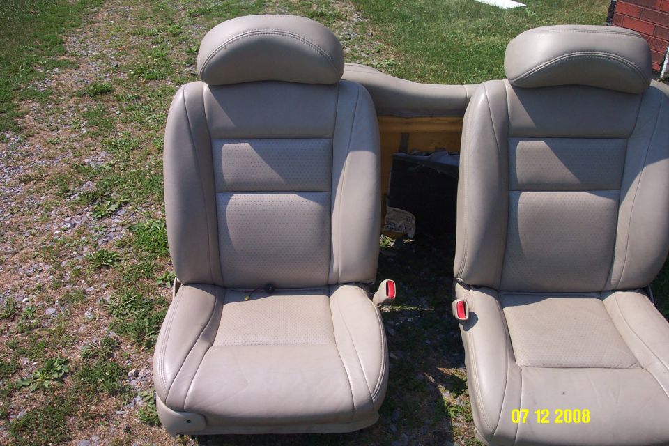 Two tan leather car seats with headrests and seatbelt buckles visible, placed outdoors on grass and gravel